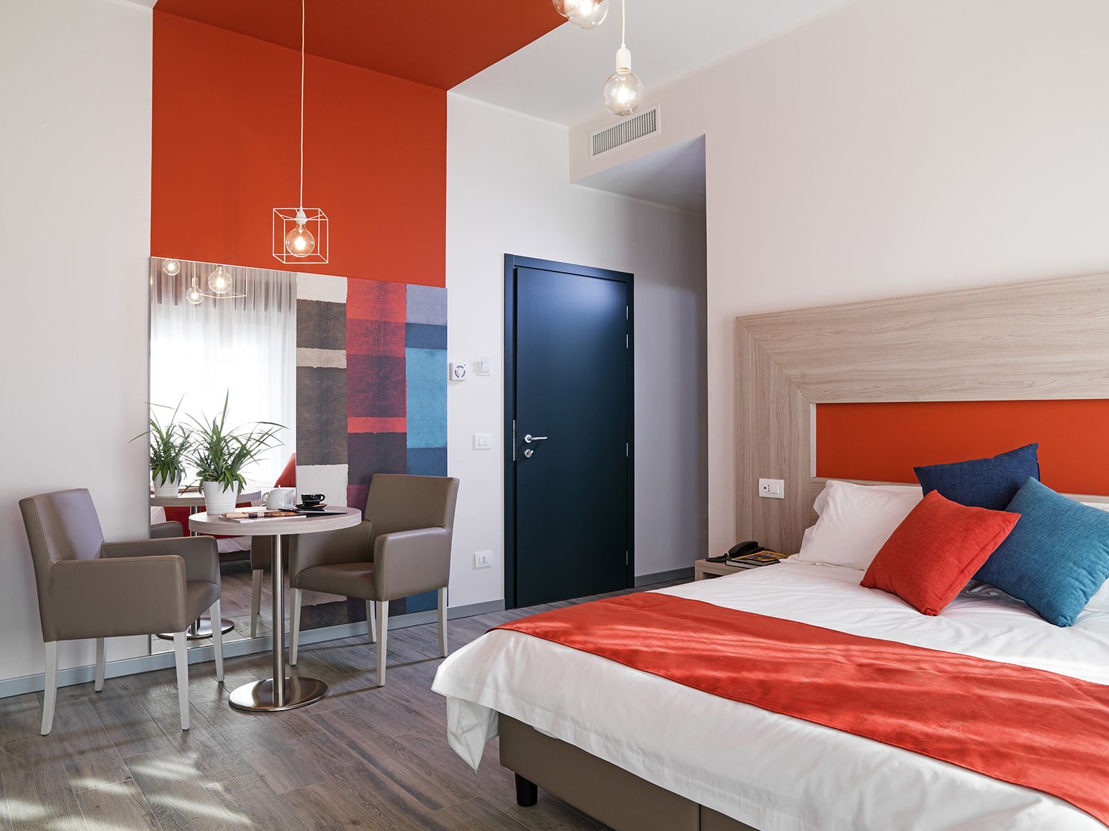 A modern hotel room with a large bed, a dining area with a table and chairs, and a vibrant abstract wall.