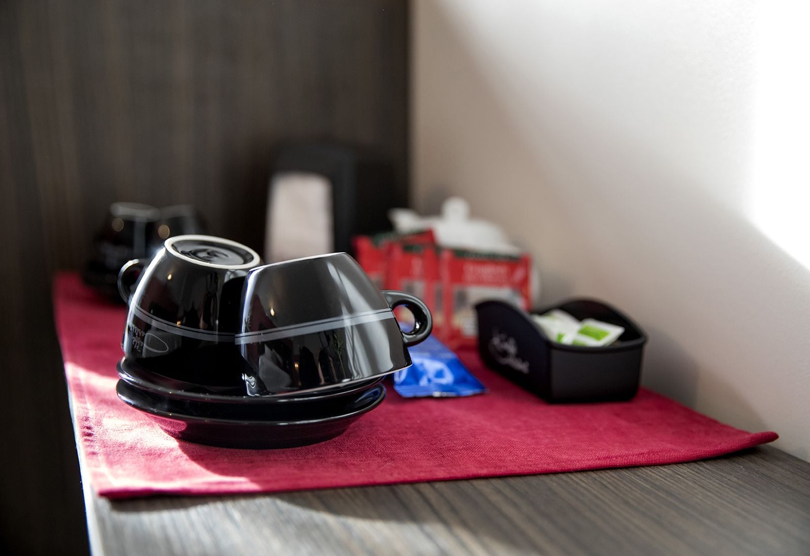 A coffee maker and a tea kettle on a wooden table with a red cloth, and a box of coffee pods in the background.