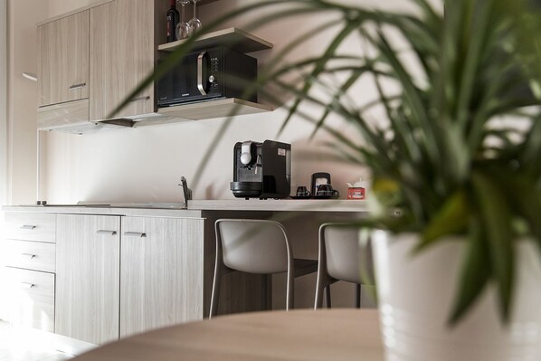 A modern kitchen with a built-in microwave, coffee maker, and wine glasses on a shelf. There are two chairs and a potted plant in the foreground.