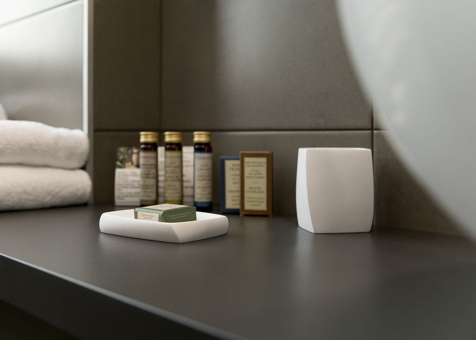 A bathroom counter with a white soap dish, a white candle, and several bottles of toiletries.