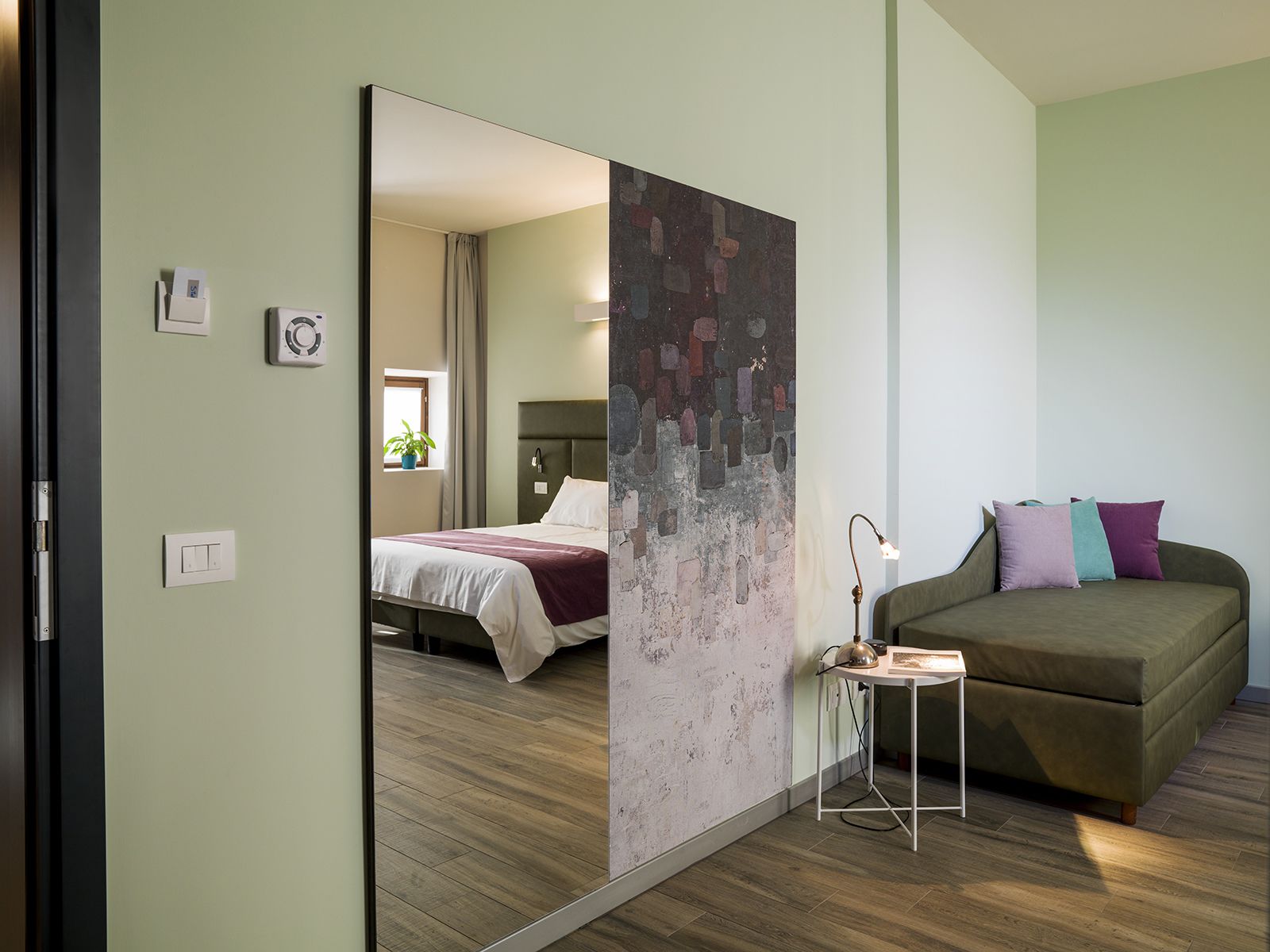 A modern bedroom with a large mirror, a bed with a headboard, a bedside table with a lamp, and a green sofa.