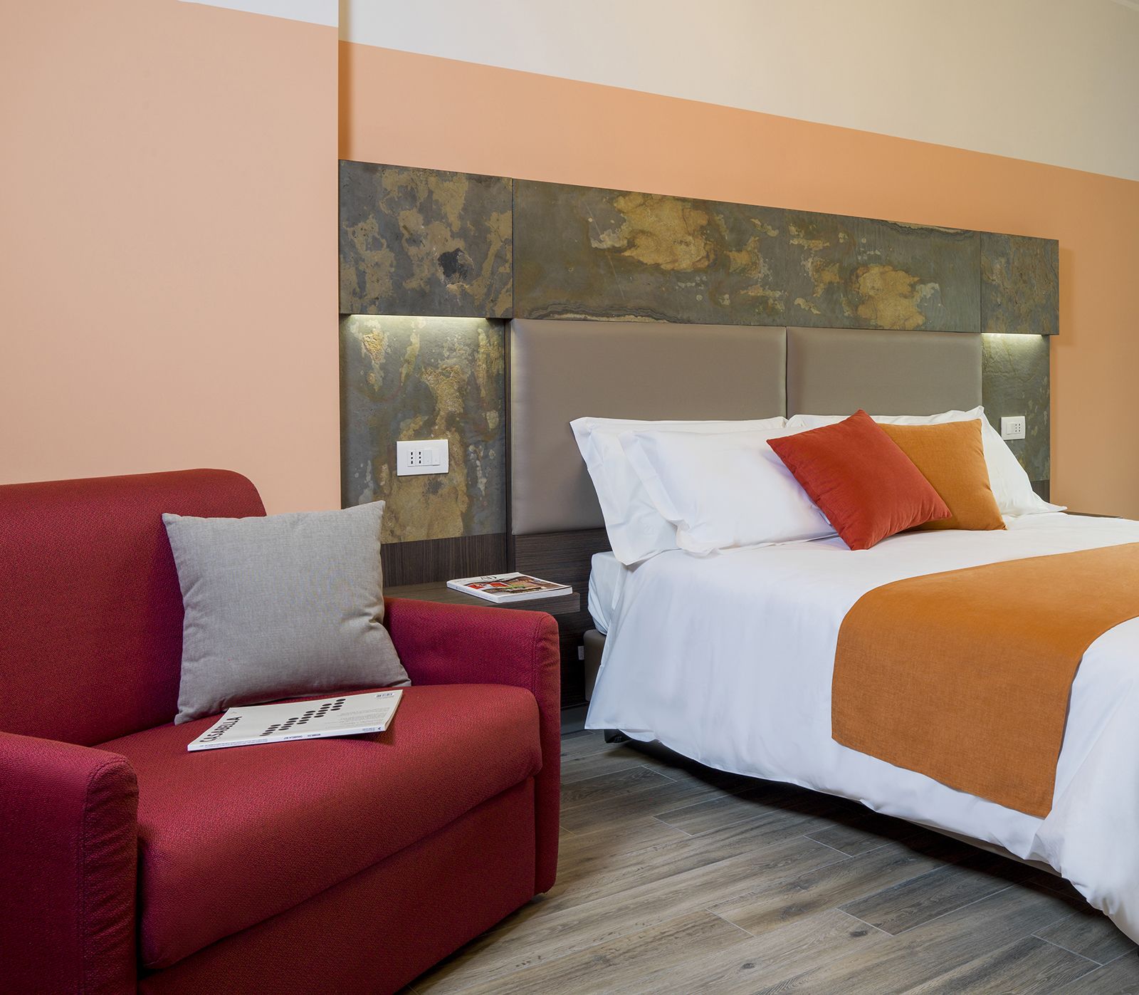Une chambre d’hôtel avec un canapé rouge, un lit avec une literie blanche et orange, et un mur texturé au-dessus du lit.