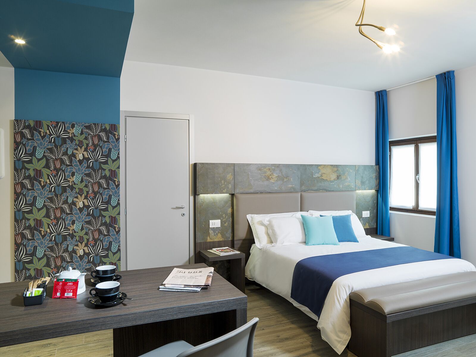 A modern hotel room with a bed, bedside table, and a desk with a chair. There is a window with curtains and a wall with a patterned wallpaper.