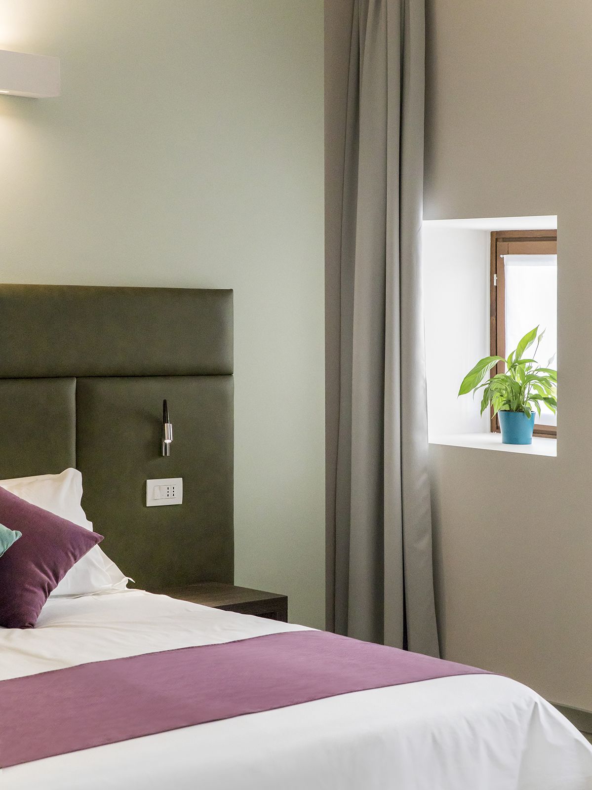 A neatly made bed with a headboard, a bedside table with a lamp, a power outlet, and a window with a plant.