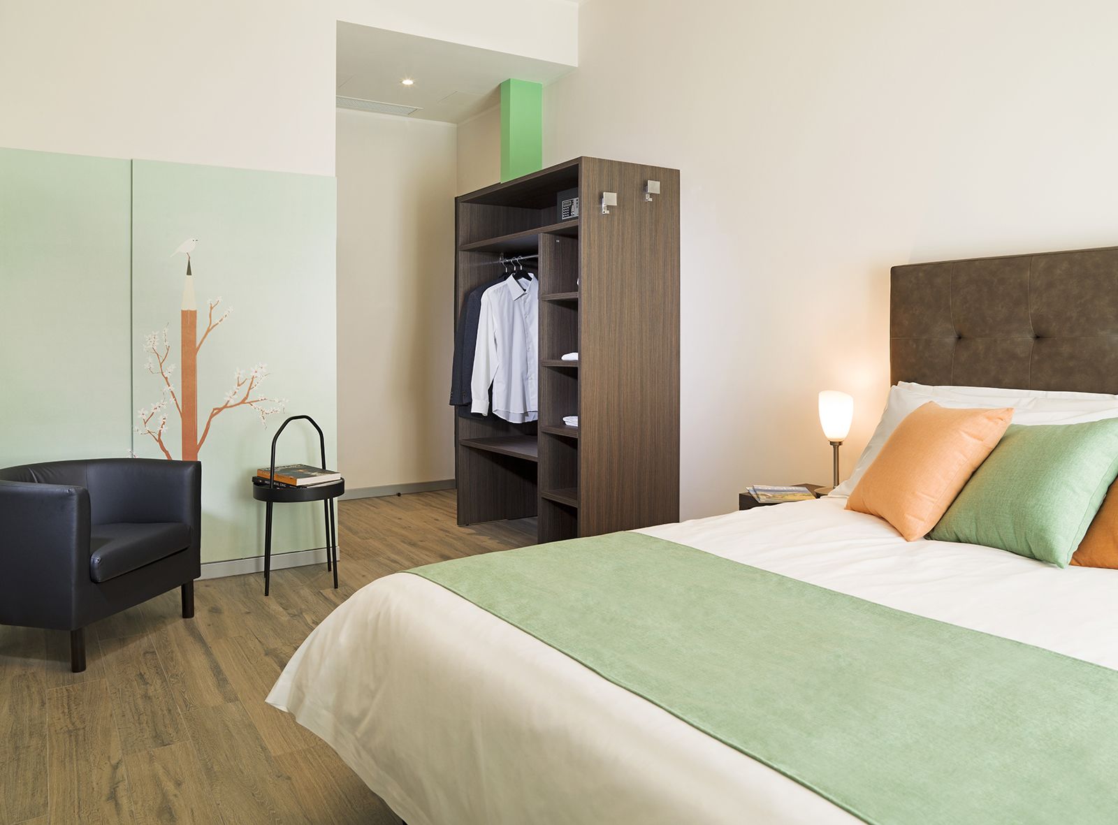 A hotel room with a bed, a chair, a wardrobe, and a nightstand with a lamp.
