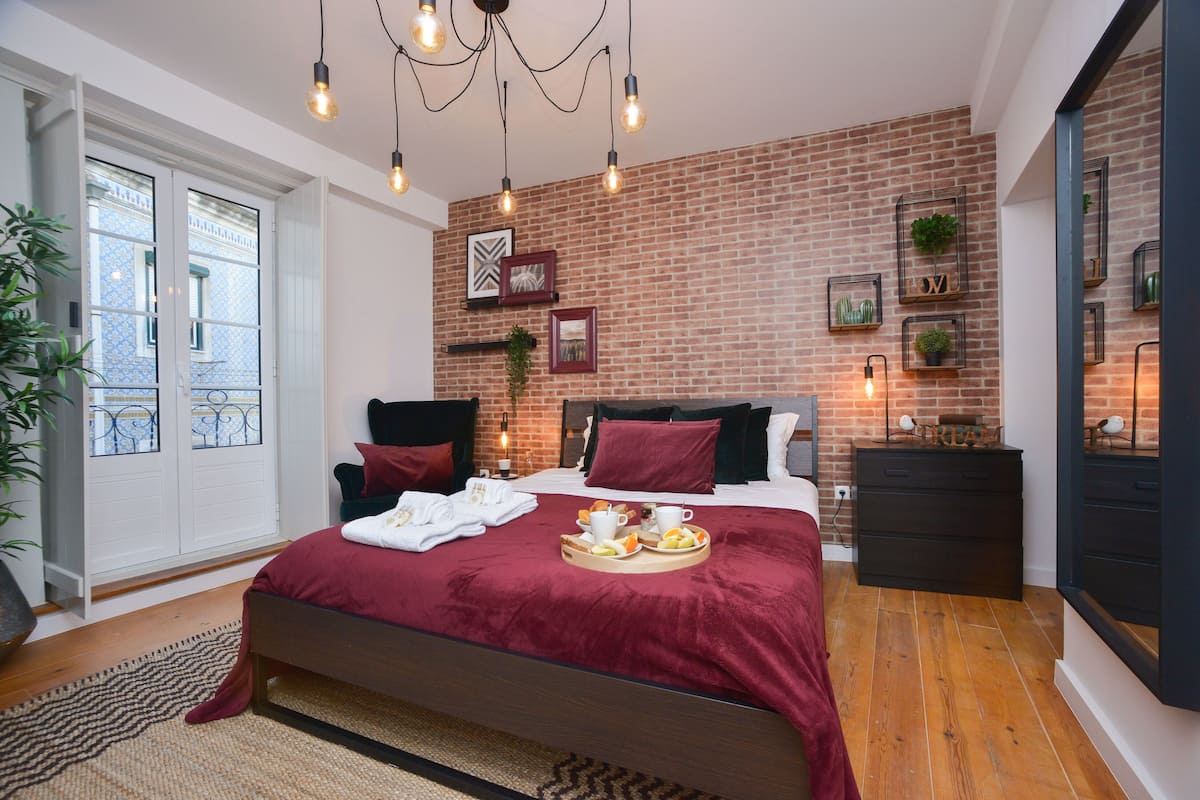 Ein modernes Schlafzimmer mit Backsteinwand, einem großen Bett mit roter Decke, Nachttischen und Blick auf einen Balkon.