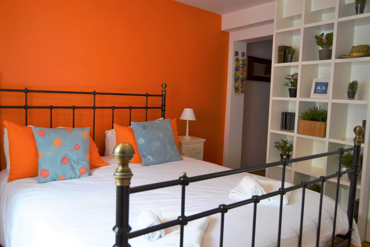 Ein Bett mit Metallgestell, weißer Bettwäsche und orange-blauen Kissen. Ein weißer Nachttisch mit Lampe und Ablagefläche mit Pflanzen und Büchern.