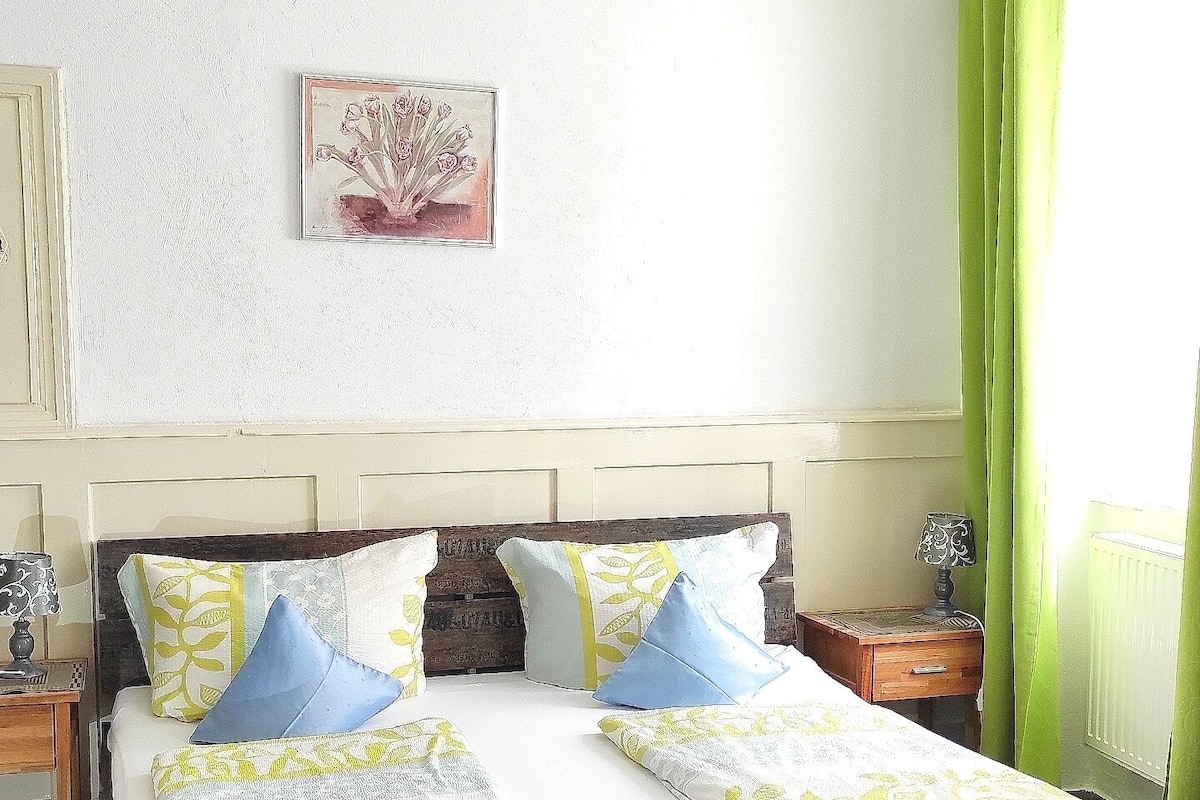 Una habitación con una cama de madera, cortinas verdes, una mesita de noche, una lámpara y un aplique de pared.