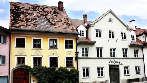 A row of traditional European buildings with distinct architectural styles, including a yellow building with arched windows and a white building with a sign reading "MangMaurus". - Maurushaus (Fuessen)