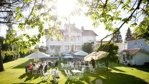 A spacious outdoor patio with multiple tables and chairs, surrounded by lush greenery and a large building in the background.