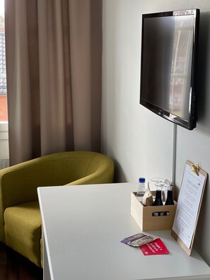 A hotel room with a flat-screen TV mounted on the wall, a white table with a clipboard and a bottle of water, and a mustard-colored armchair.