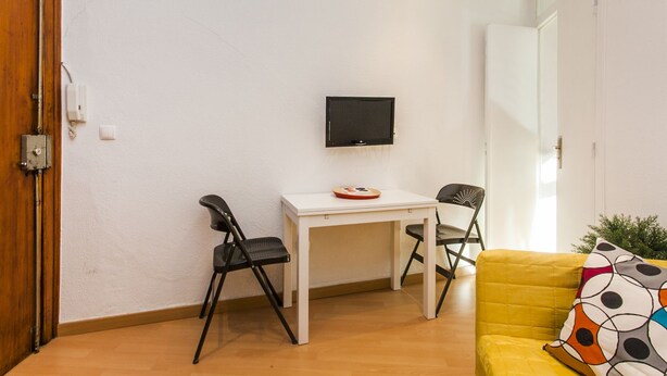 Une petite pièce lumineuse, avec une table blanche, deux chaises noires, un canapé jaune et un téléviseur fixé au mur.