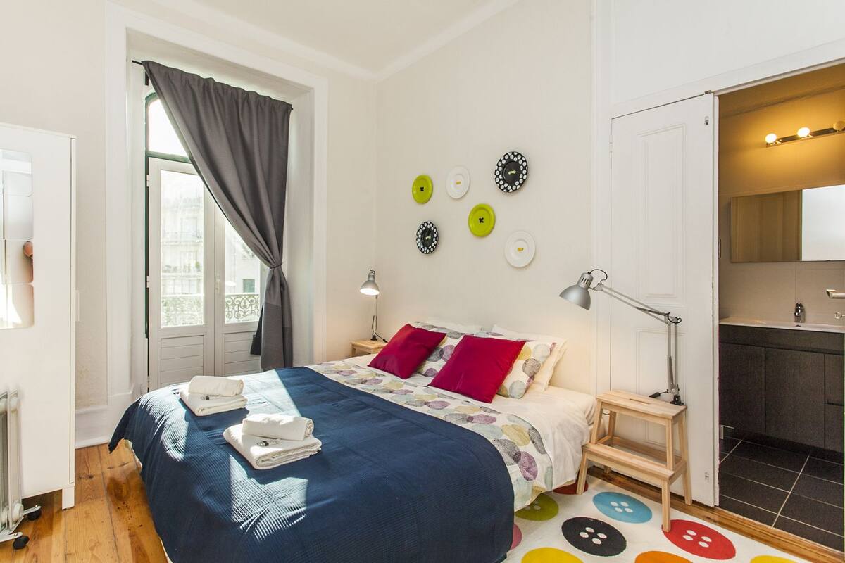 Un dormitorio ordenado con una cama, mesitas de noche, una lámpara y vistas a la ciudad a través de una ventana.