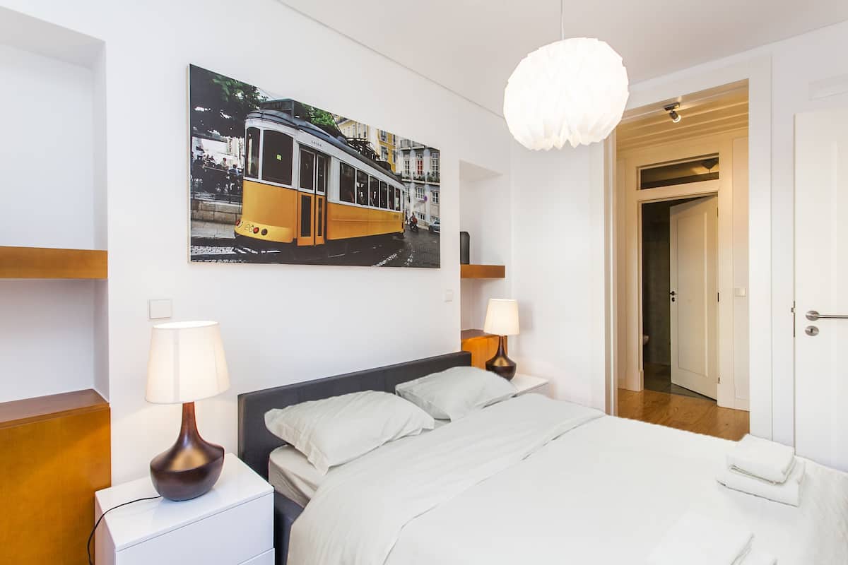 A hotel room with a bed, two bedside lamps, a painting of a tram, and a door leading to a bathroom.