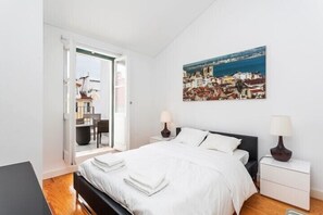 A bedroom with a bed, bedside tables, a painting of a coastal town, and a view through an open door.