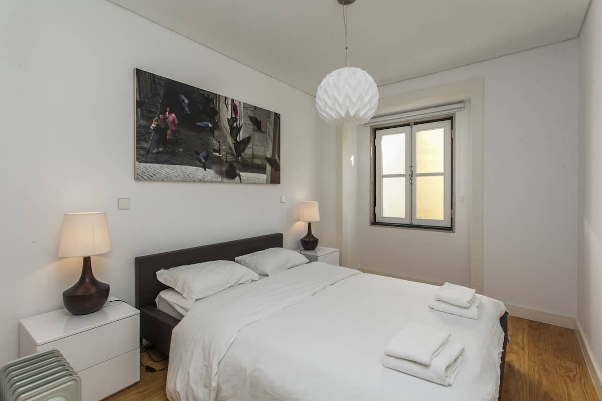 A neatly arranged bedroom with a bed, two bedside tables with lamps, a large window, and a wall-mounted artwork.
