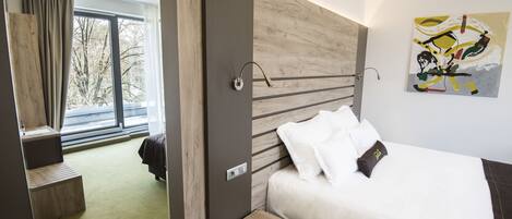 Habitación de hotel moderna con una cama grande, una mesita de noche pequeña, una ventana con cortinas y vistas a árboles.