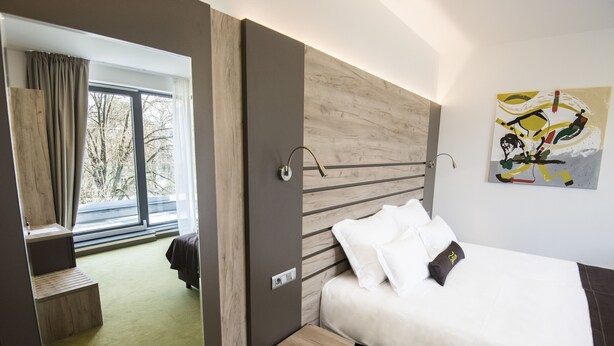 Habitación de hotel moderna con una cama grande, una mesita de noche pequeña, una ventana con cortinas y vistas a árboles.