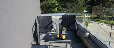 A balcony with two wicker chairs, a small table with a fruit bowl and two glasses of wine, and a potted plant.