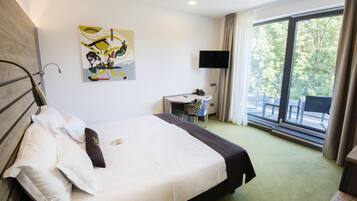 Habitación de hotel con cama, escritorio, silla, televisor y ventana con vista a árboles.