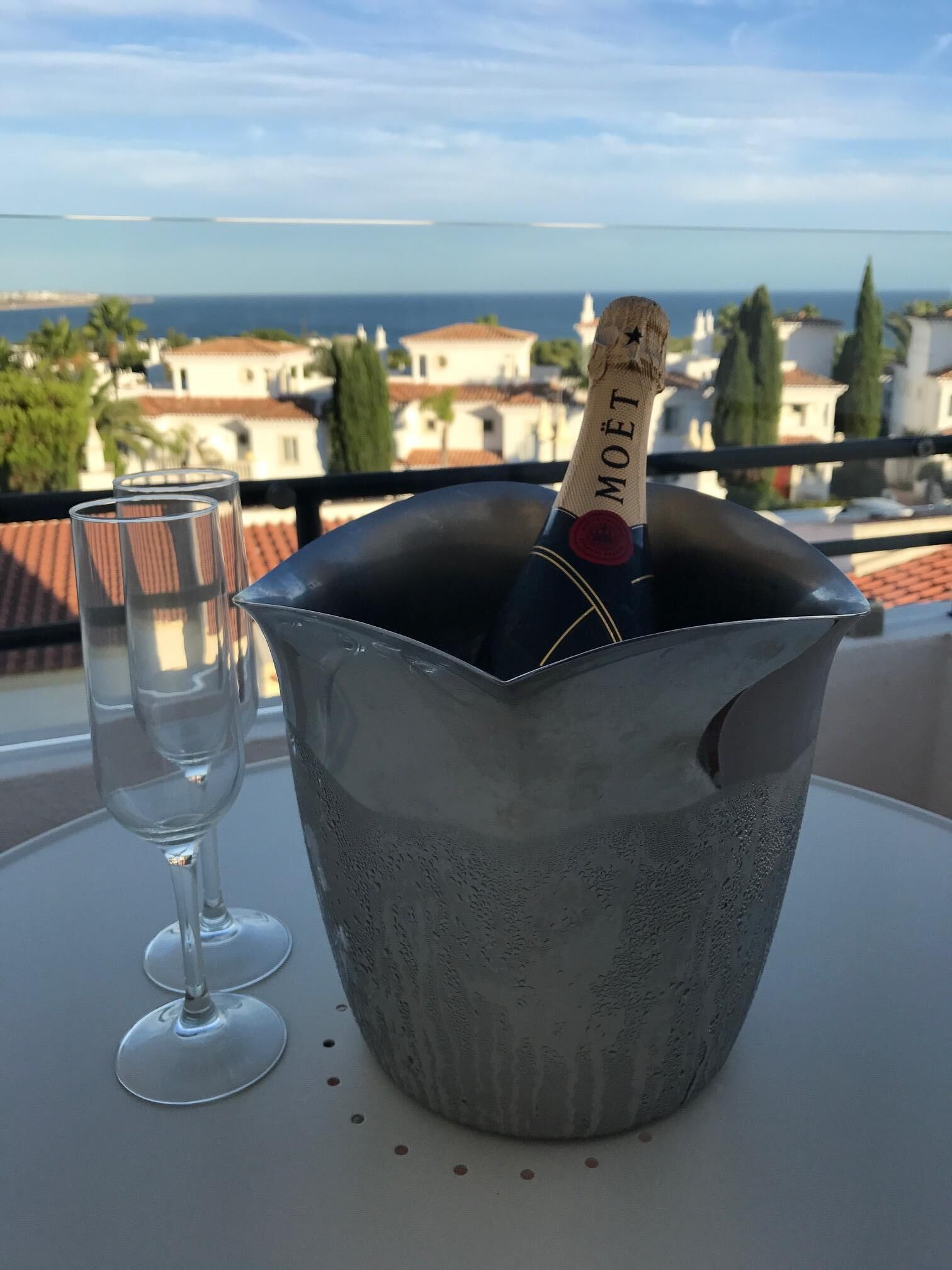 A metallic champagne bucket with a bottle labeled "MOËT" on a balcony table, accompanied by two empty wine glasses.