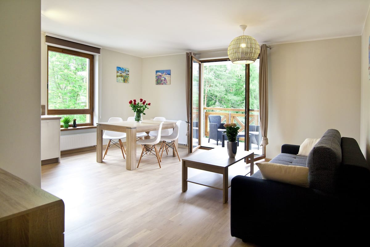 A modern living room with a dining area, a sofa, a coffee table, and a view of a balcony with outdoor furniture.