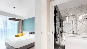 A modern hotel room with a large bed, a bright window, and a minimalist bathroom with a glass partition.