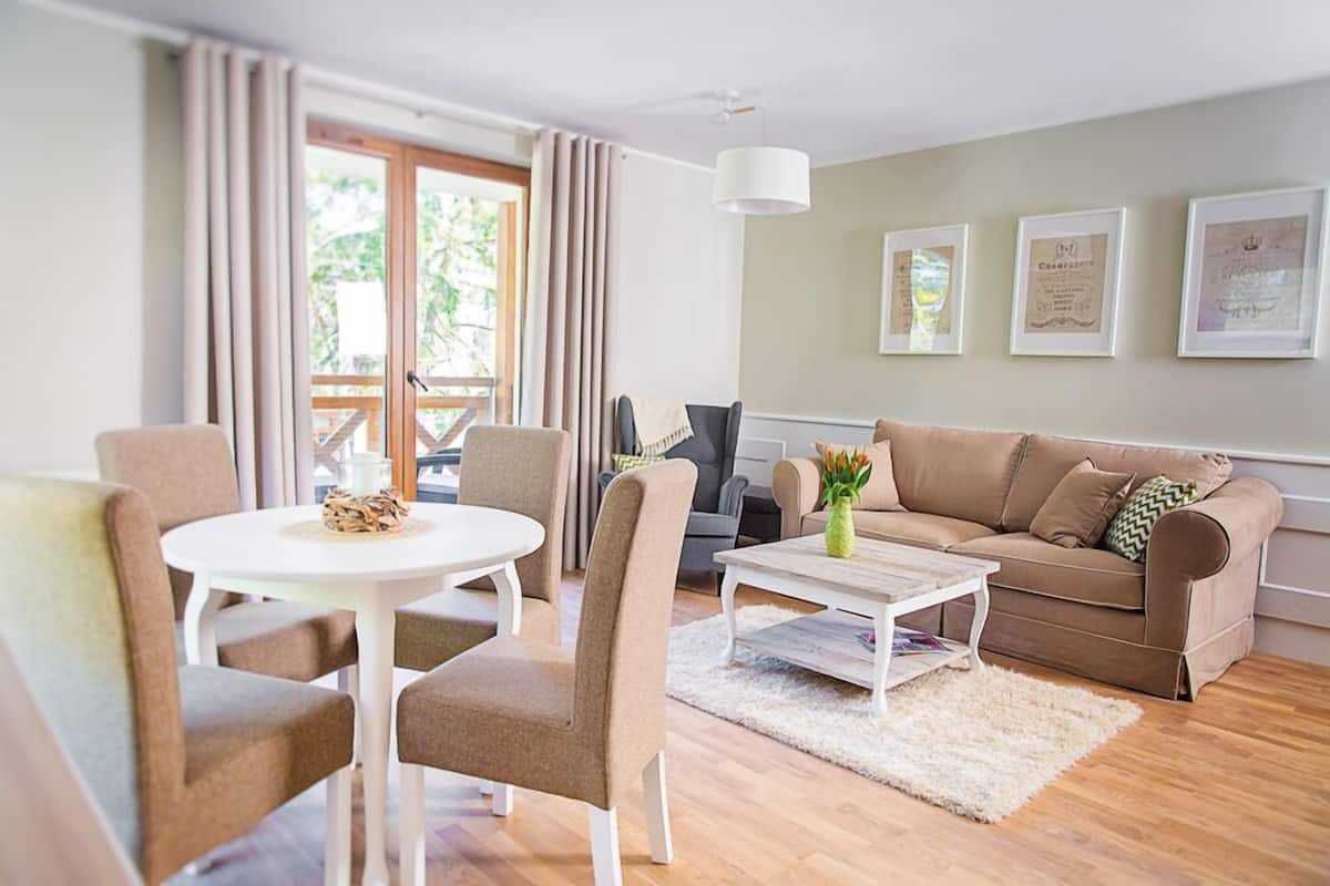 A cozy living room with a white round table, beige sofa, and a coffee table. There are framed certificates on the wall and a large window with curtains.