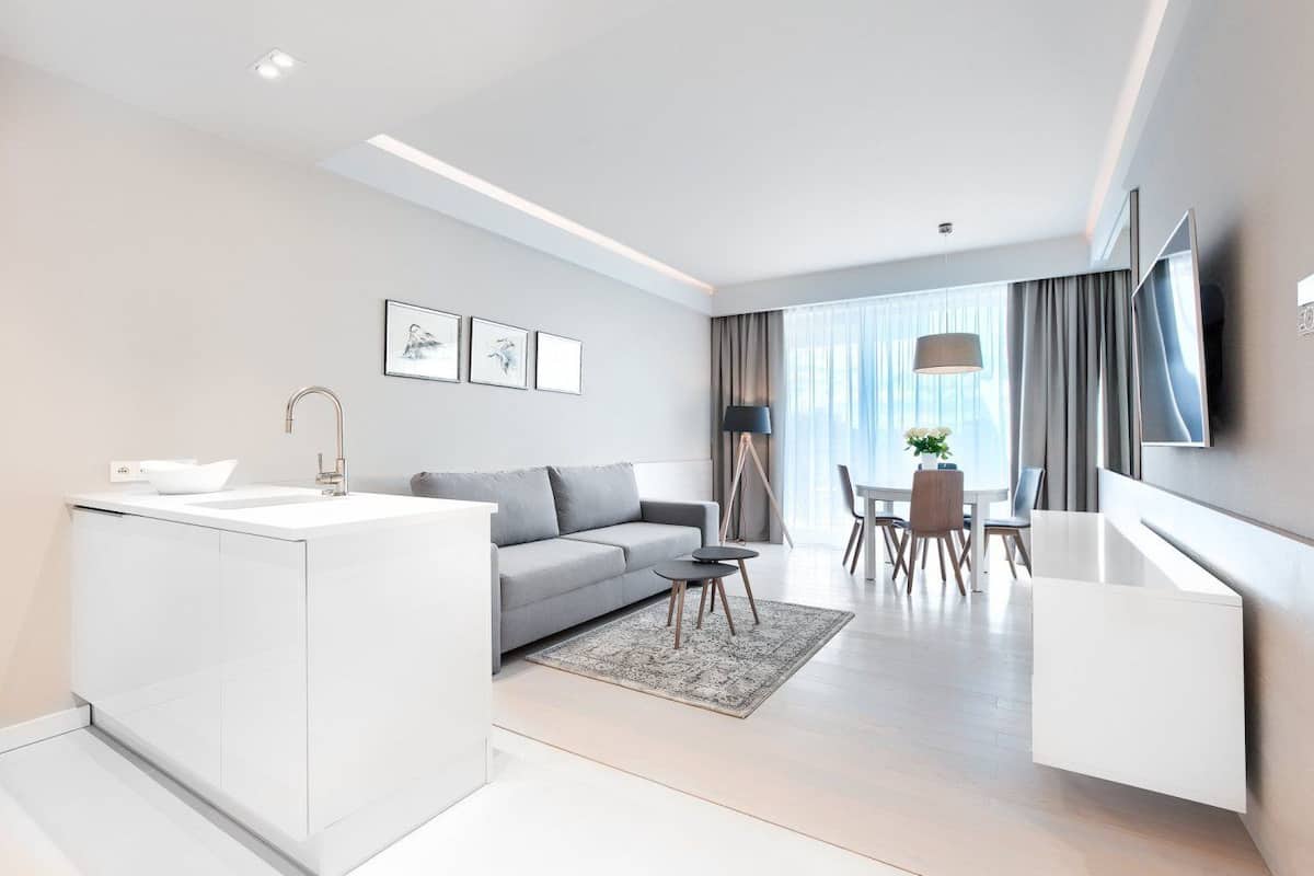A modern, well-lit living area with a kitchen island, grey sofa, dining table, and a TV mounted on the wall.