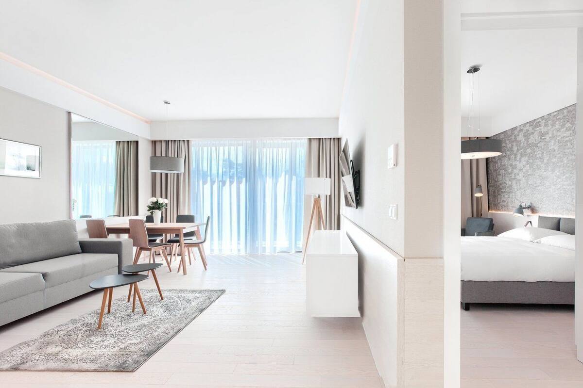 A modern hotel room with a grey sofa, a dining area with a table and chairs, a large window with curtains, and a bed with a grey headboard.