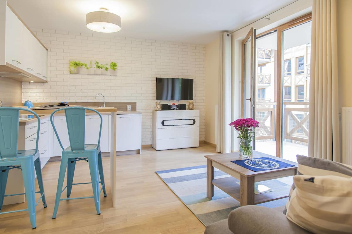 A modern kitchen with white cabinets, a dining area with blue chairs, a flat-screen TV above the counter, and a sliding glass door leading to a balcony.