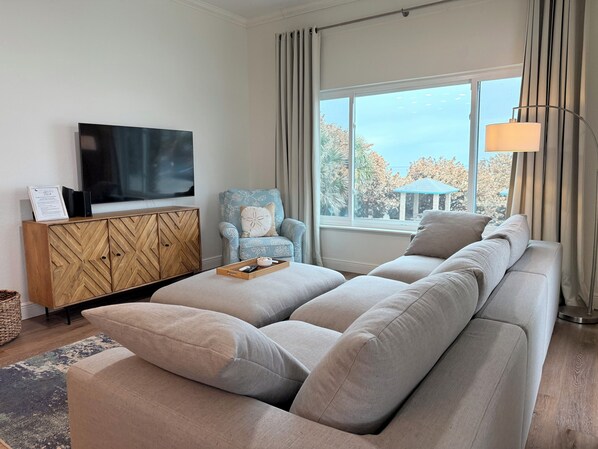Una sala de estar moderna con un sofá seccional grande, un mueble de madera con un televisor y vistas a los árboles a través de amplios ventanales.