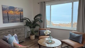 Una sala de estar con vista a la playa, un cuadro con una hamaca, una planta grande y una mesa de centro redonda con un jarrón.