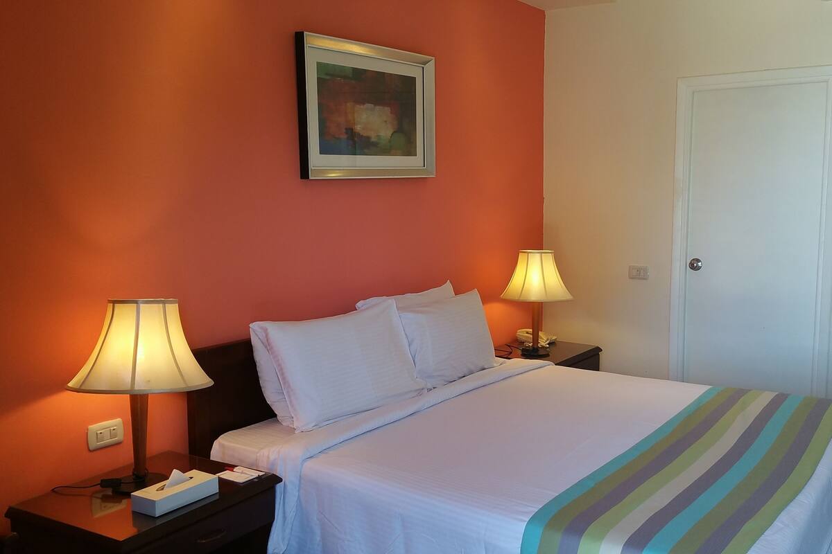 Una habitación de hotel con una cama, dos lámparas de noche, un cuadro en la pared y una puerta.