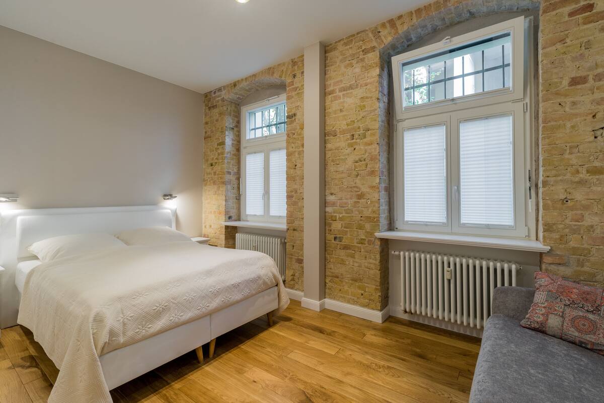 A bedroom with a bed, a grey sofa, brick walls, and large windows with blinds.