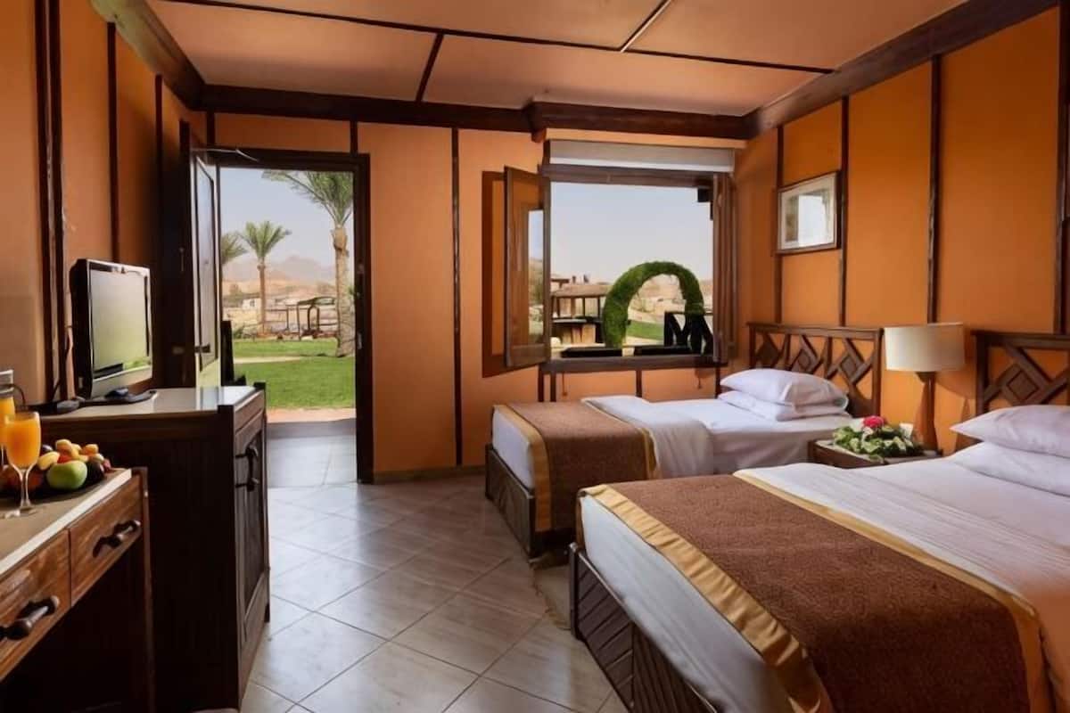 Ein Hotelzimmer mit zwei Betten, einem Schreibtisch mit Fernseher und einem Fenster mit Blick auf eine Palme und Berge.