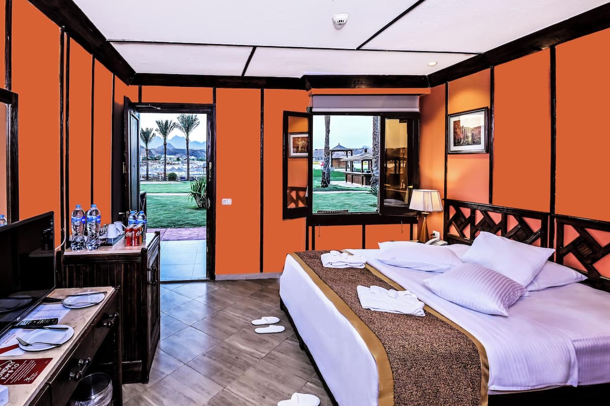 Ein Hotelzimmer mit einem Bett, einem Schreibtisch mit Computer, einem Stuhl und einem Fenster mit Blick auf Palmen und Berge.