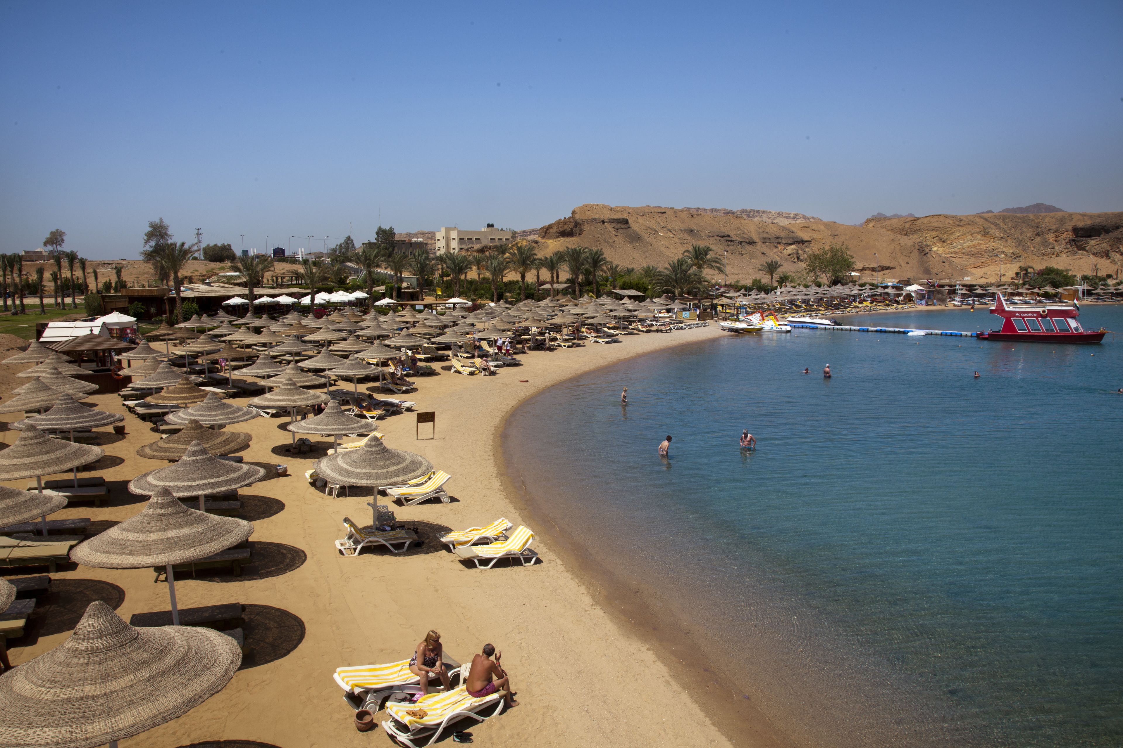 A beach with numerous sun umbrellas and lounge chairs, a clear blue sea, and a few boats.
