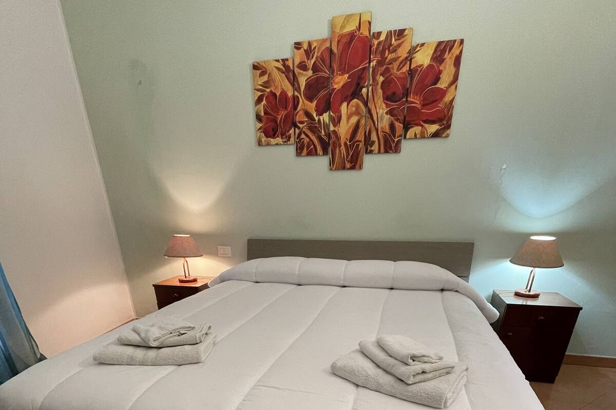 A neatly made bed with a large bedspread and two towels folded on top. There are two bedside tables with lamps, and a large floral painting on the wall.