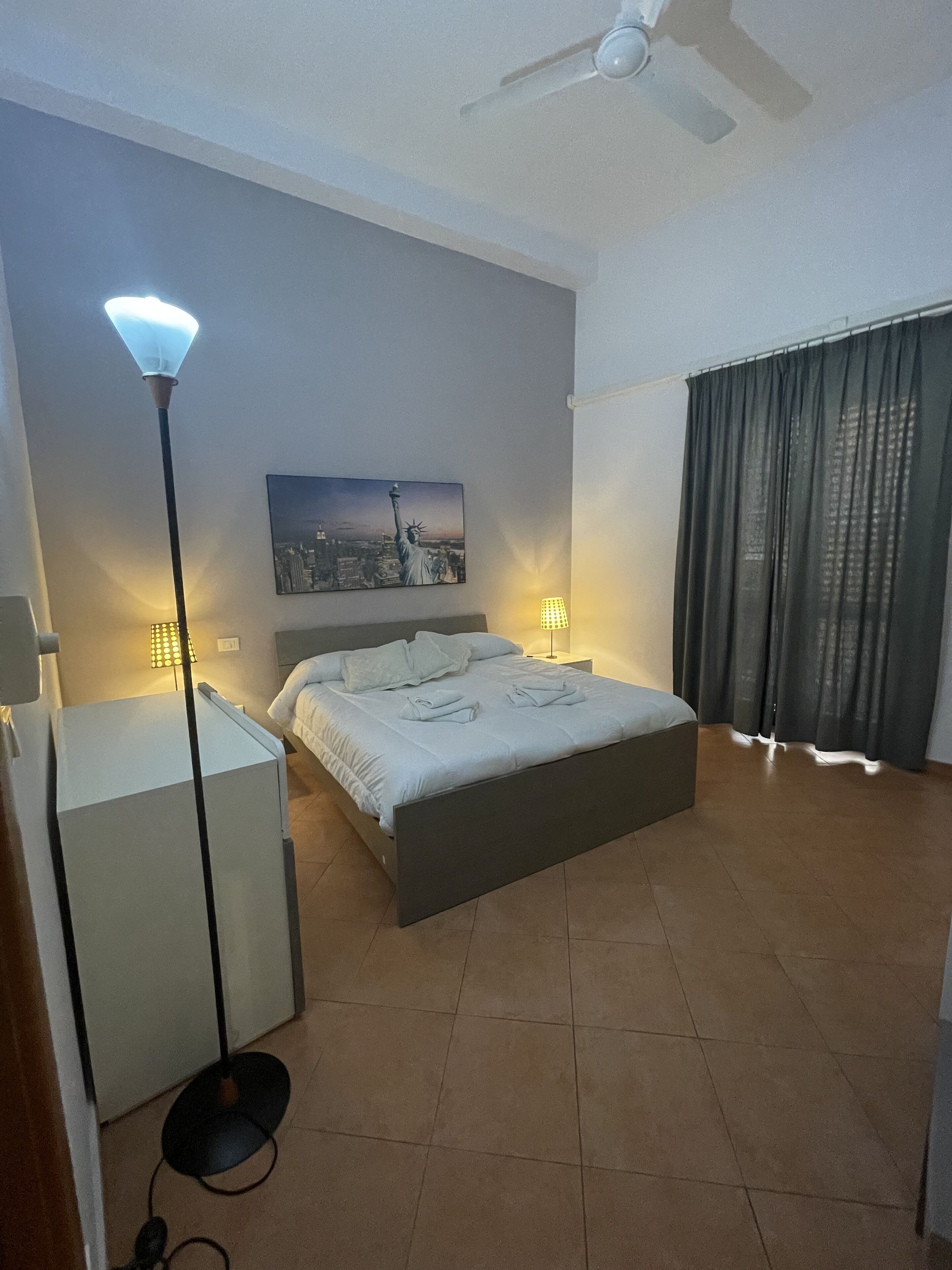 Ein modernes Schlafzimmer mit Bett, Nachttischlampen, Deckenventilator, Stadtbildgemälde und großem Fenster mit Vorhängen.