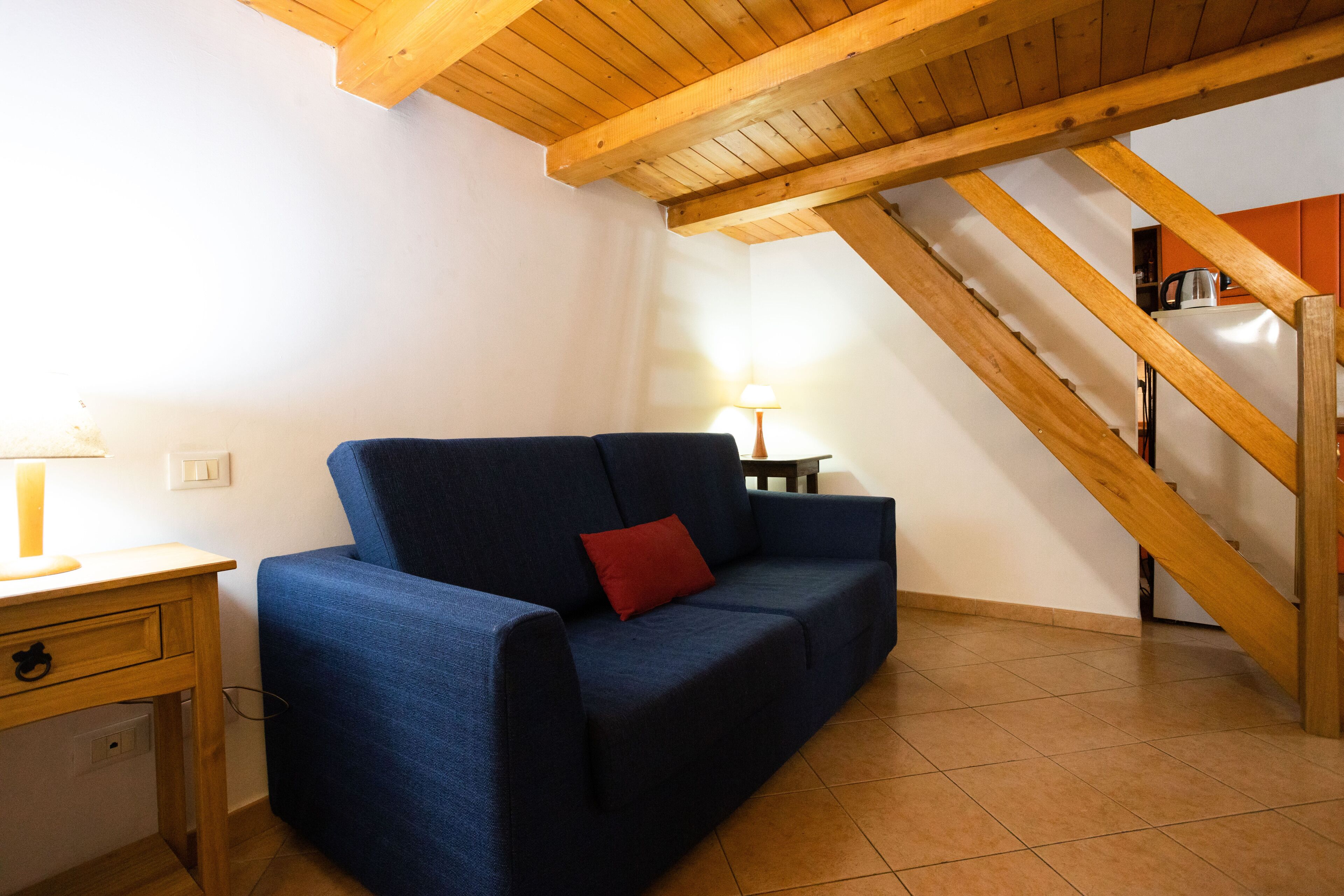 Una habitación con un sofá azul, un escritorio de madera con una lámpara y una escalera con vigas de madera.