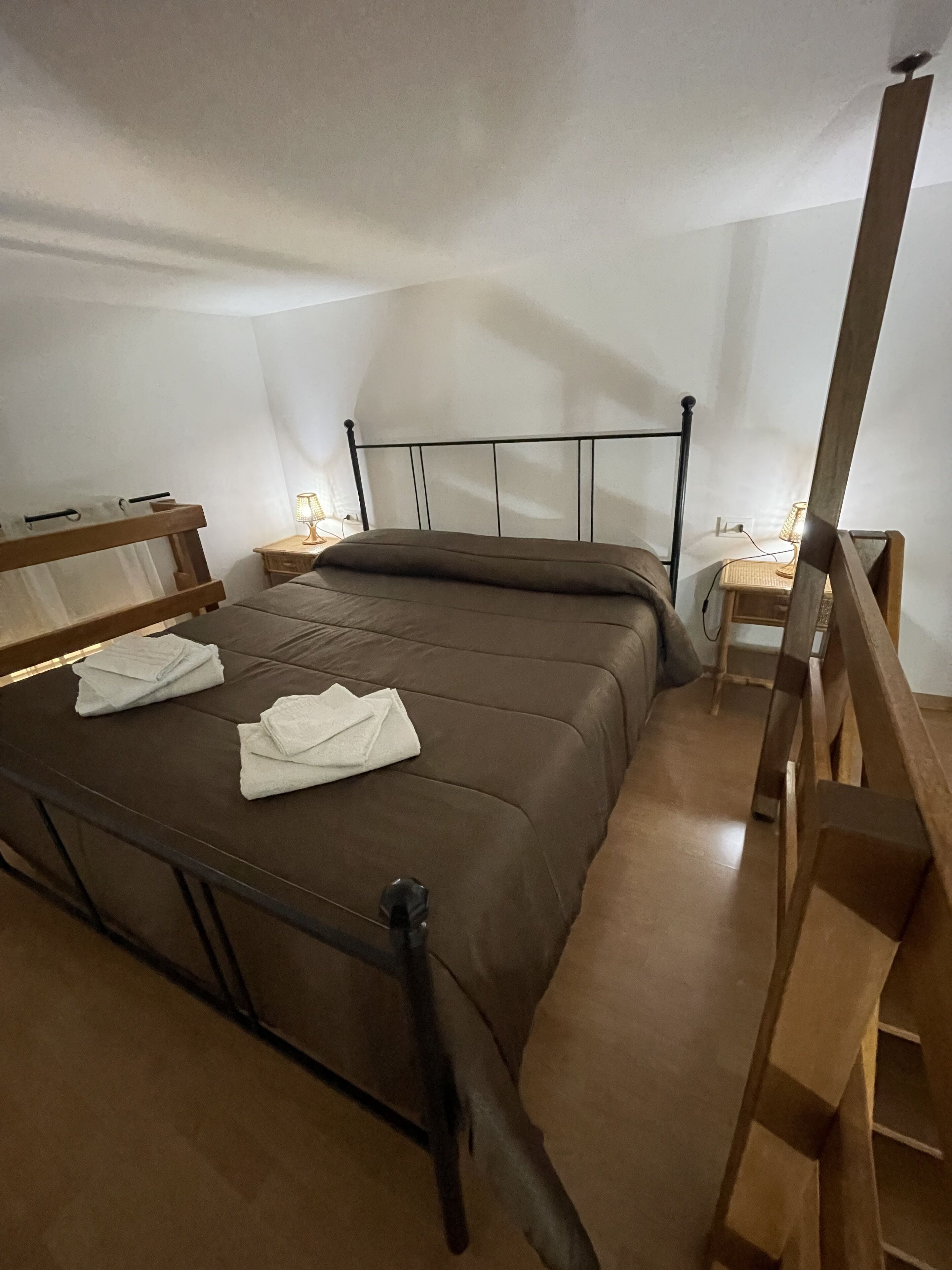 Un lit simple avec une tête de lit et un pied de lit en bois, une table de chevet avec une lampe et un petit tabouret en bois.