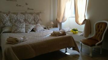 A bedroom with a bed, pillows, a chair, a nightstand, a lamp, and a window with curtains.