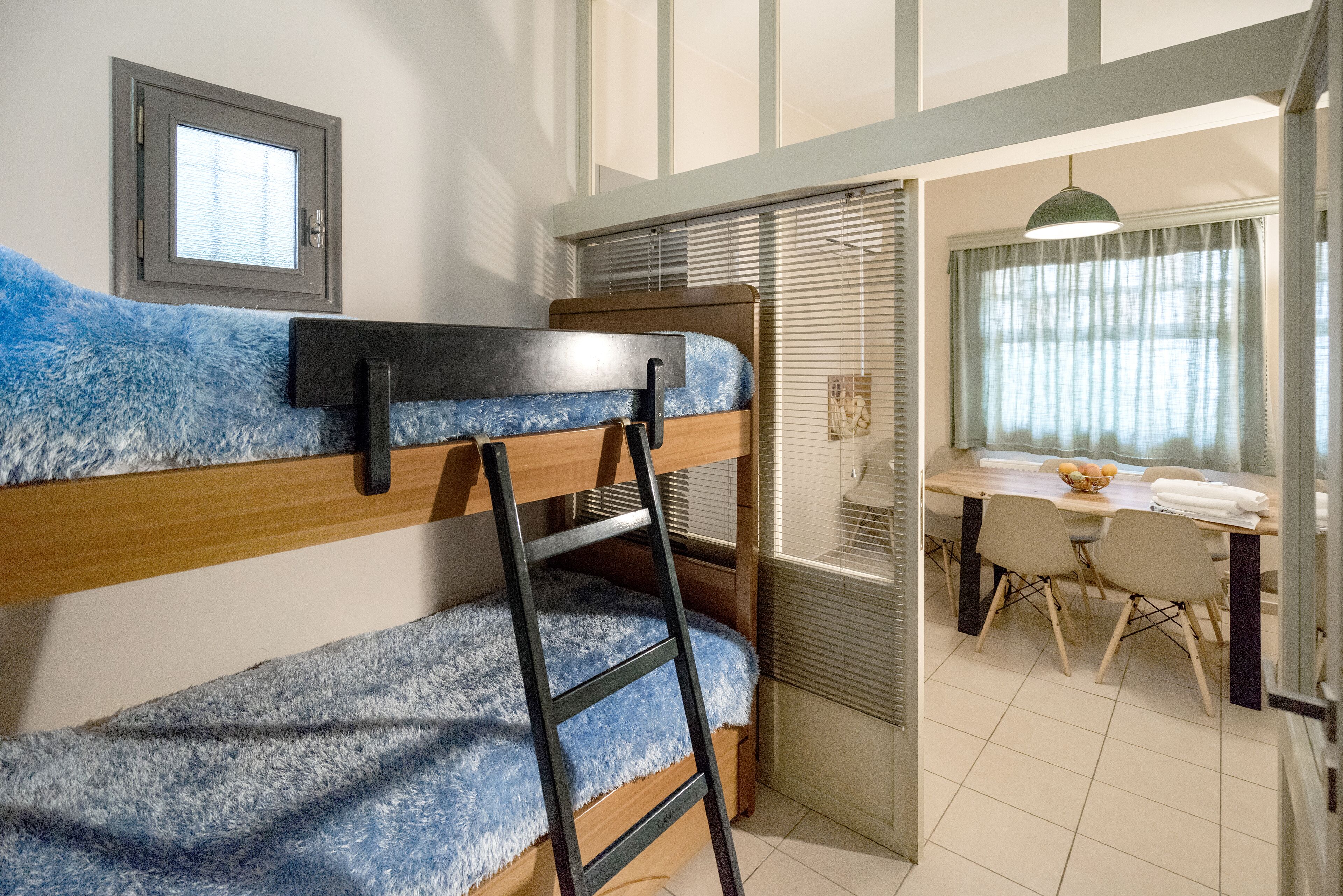 Un lit superposé avec une échelle, une fenêtre avec des stores, une table à manger avec des chaises et une suspension.