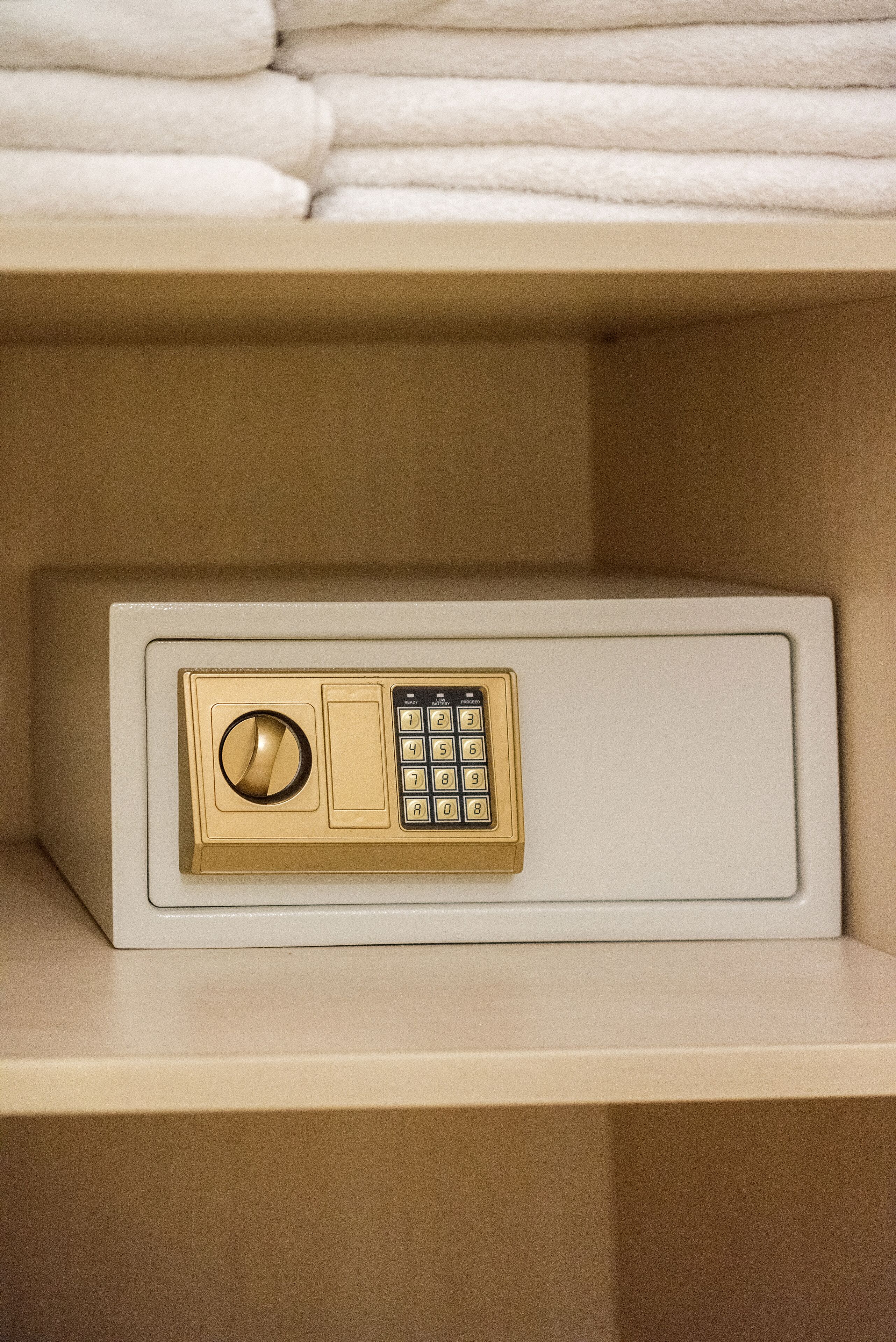 A white safe with a gold dial and keypad, located on a beige shelf above folded white towels.