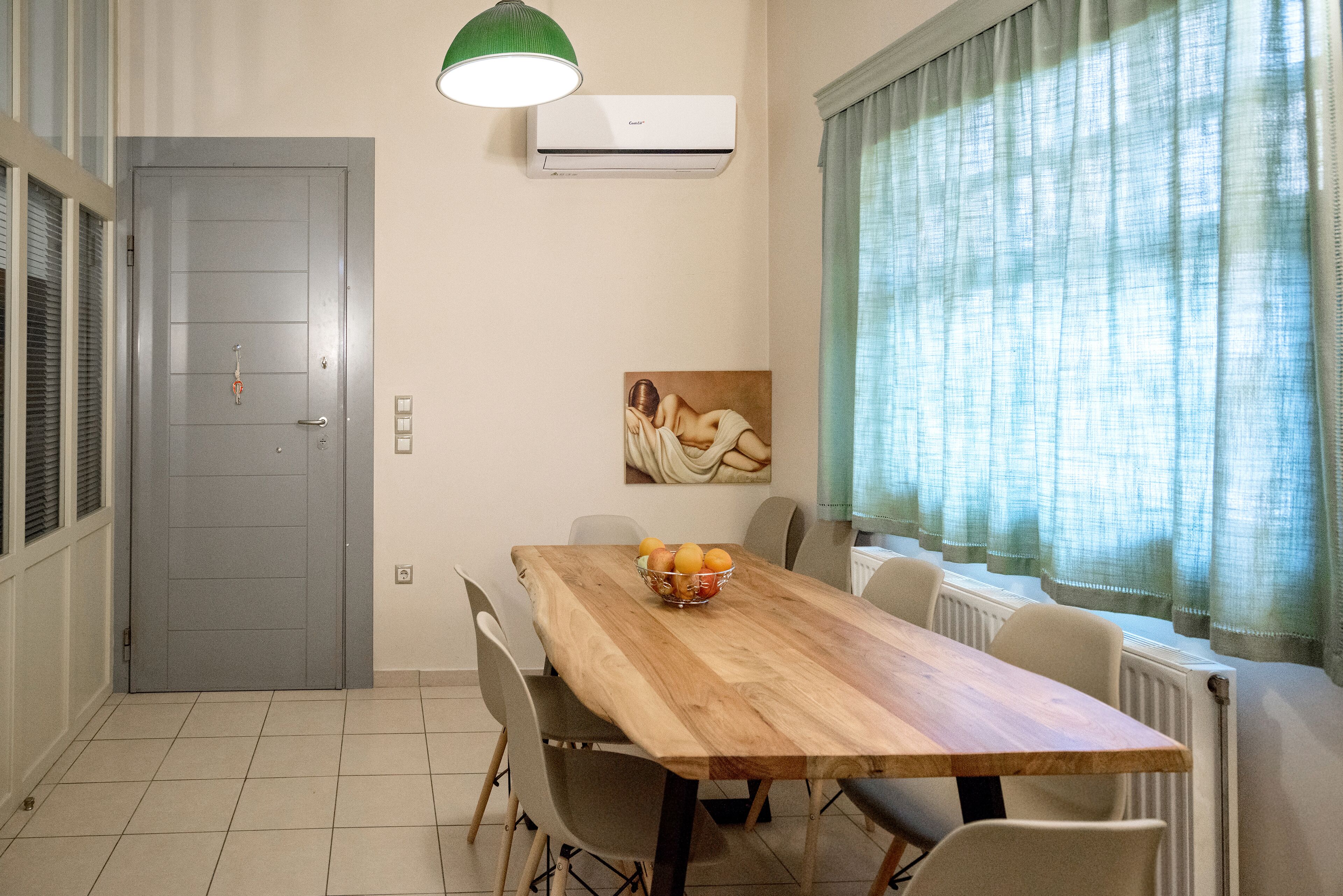 A dining area with a wooden table, chairs, a bowl of fruit, a painting on the wall, and a window with curtains.