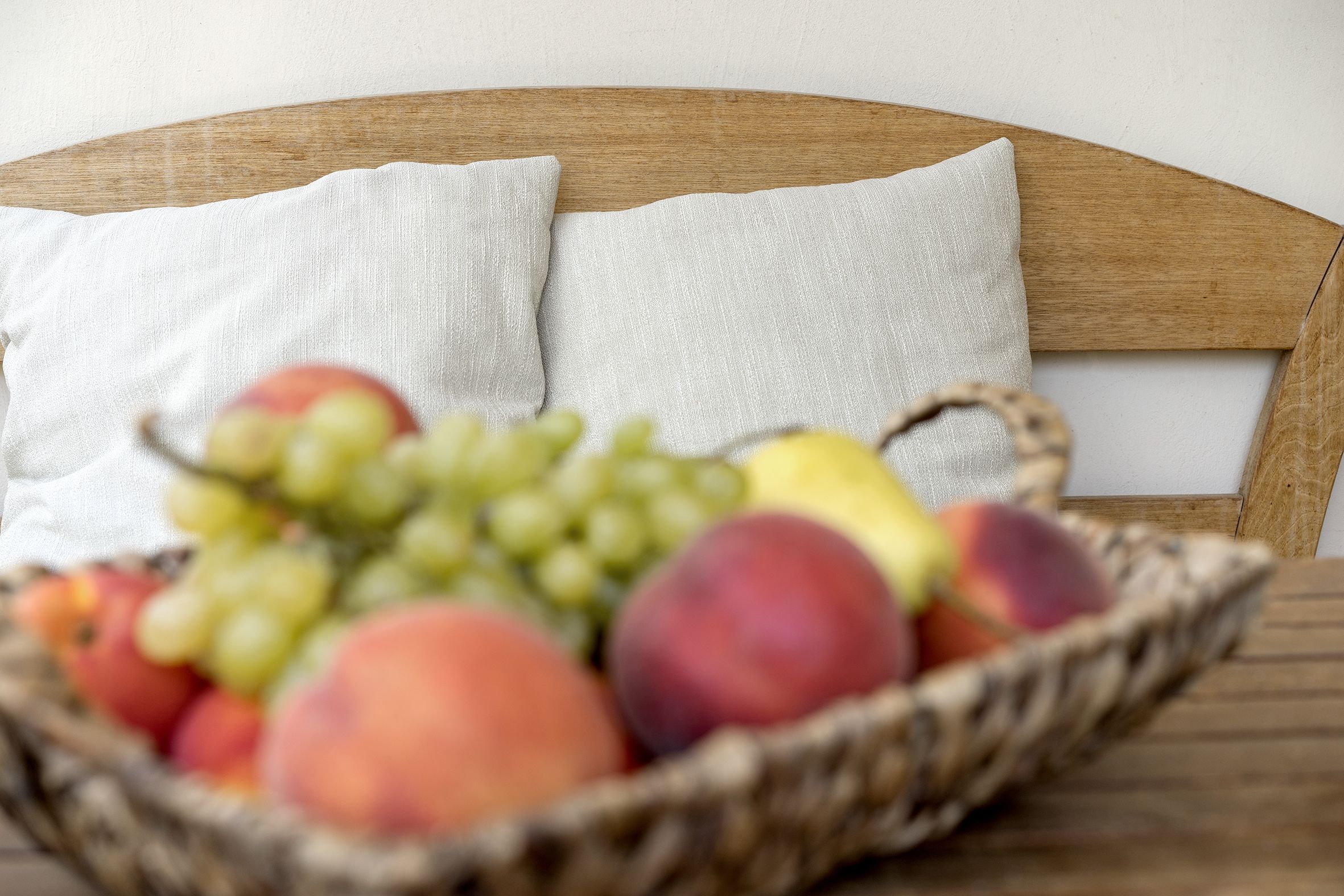 Ein Korbwesen-Korb voller Früchte auf einem Holztisch, platziert auf einem Bett mit einem hölzernen Kopfteil und zwei weißen Kissen.