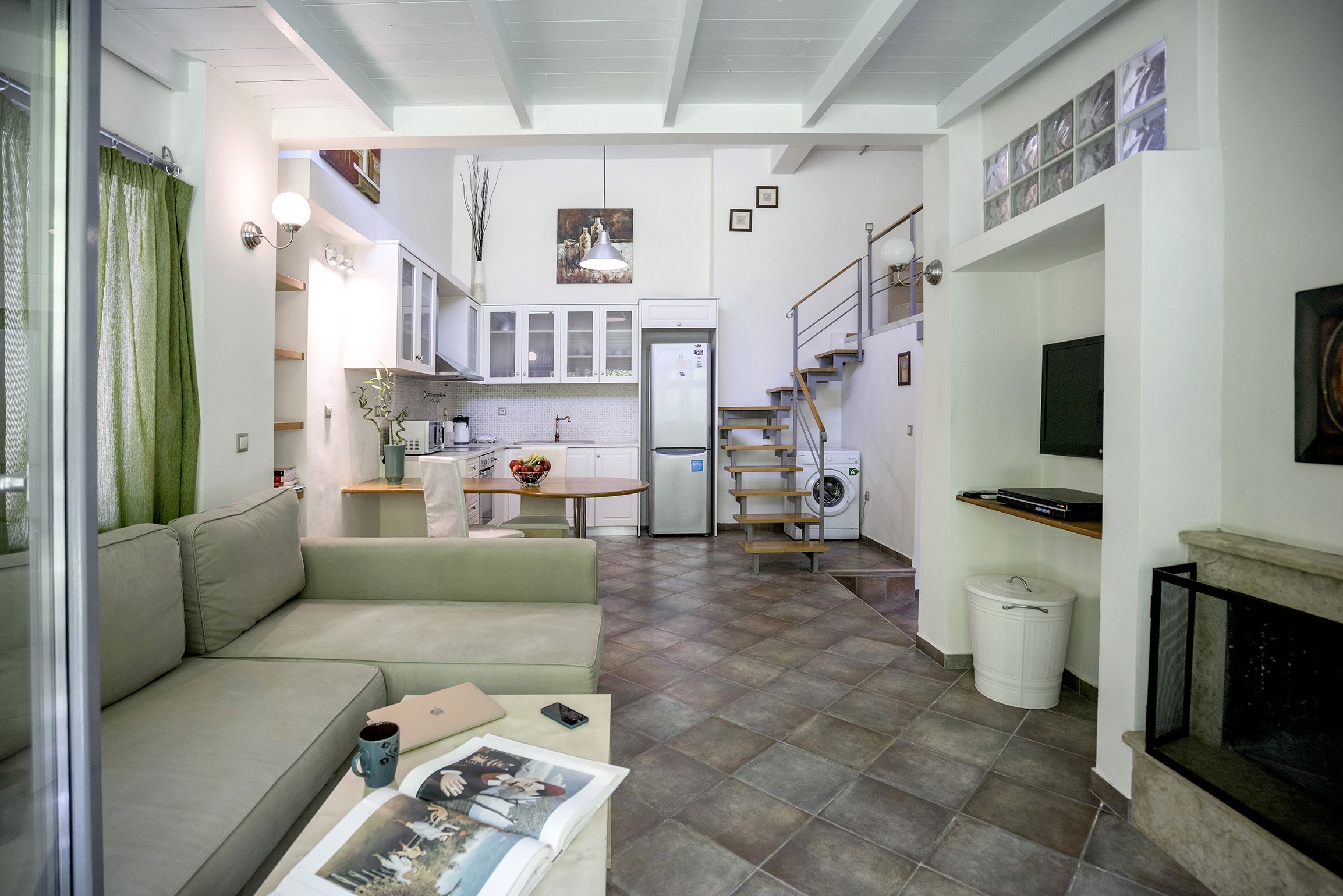 A spacious living area with a sofa, coffee table, and a fireplace. A kitchen with cabinets and a refrigerator is visible in the background.