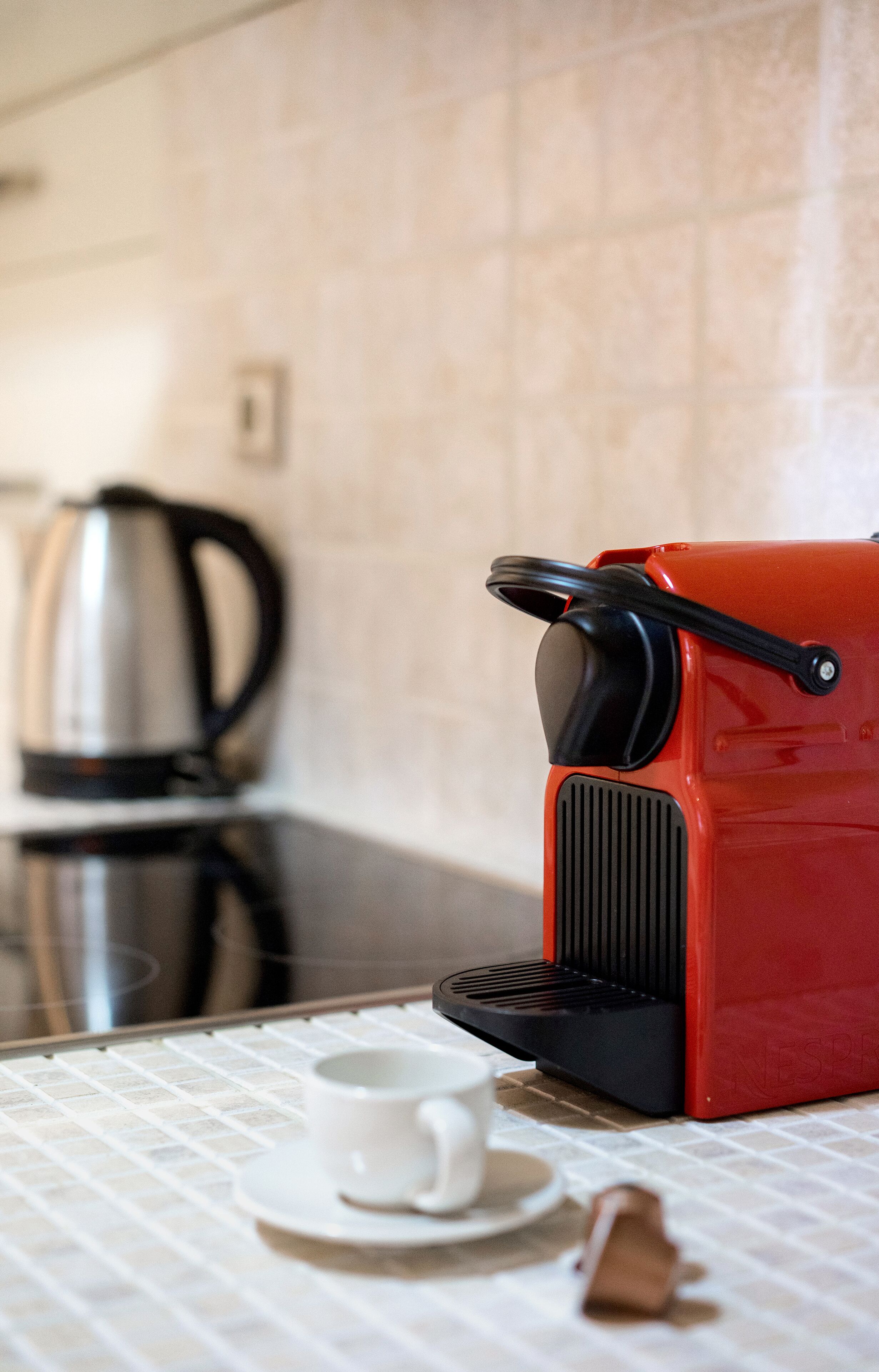 Una cocina con una cafetera roja, una taza blanca sobre un platillo y un hervidor en la encimera.