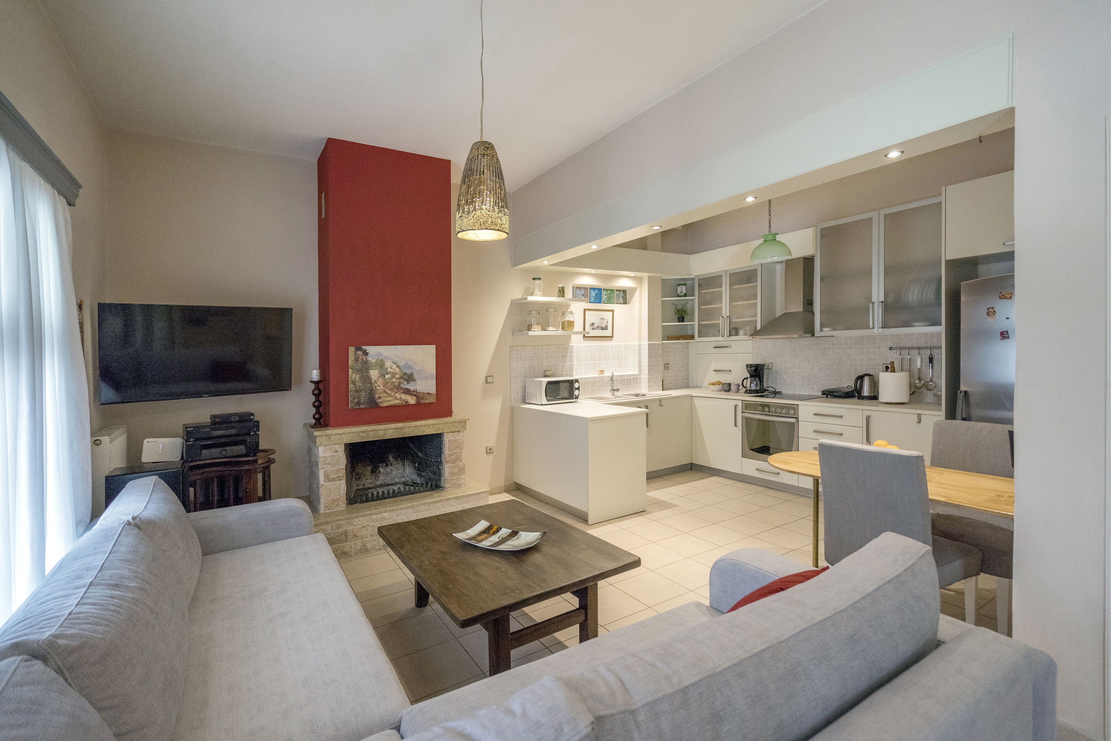 A modern living room with a sofa, a coffee table, and a TV. Adjacent is a kitchen with white cabinets, a dining area, and a fireplace.